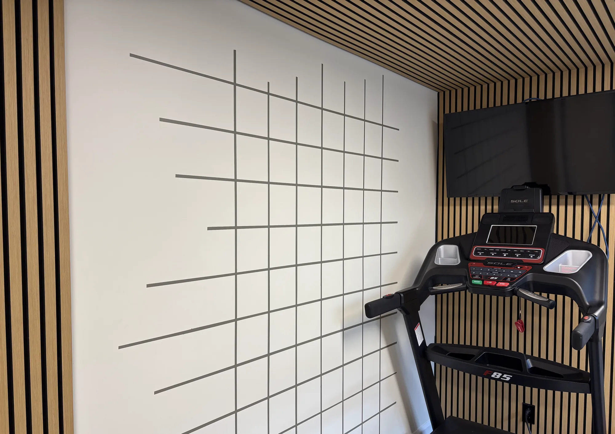 Salle d'analyse : mur crème quadrillé de lignes grises, lattes de bois au plafond et sur le mur de gauche, tapis roulant noir et écran mural à droite