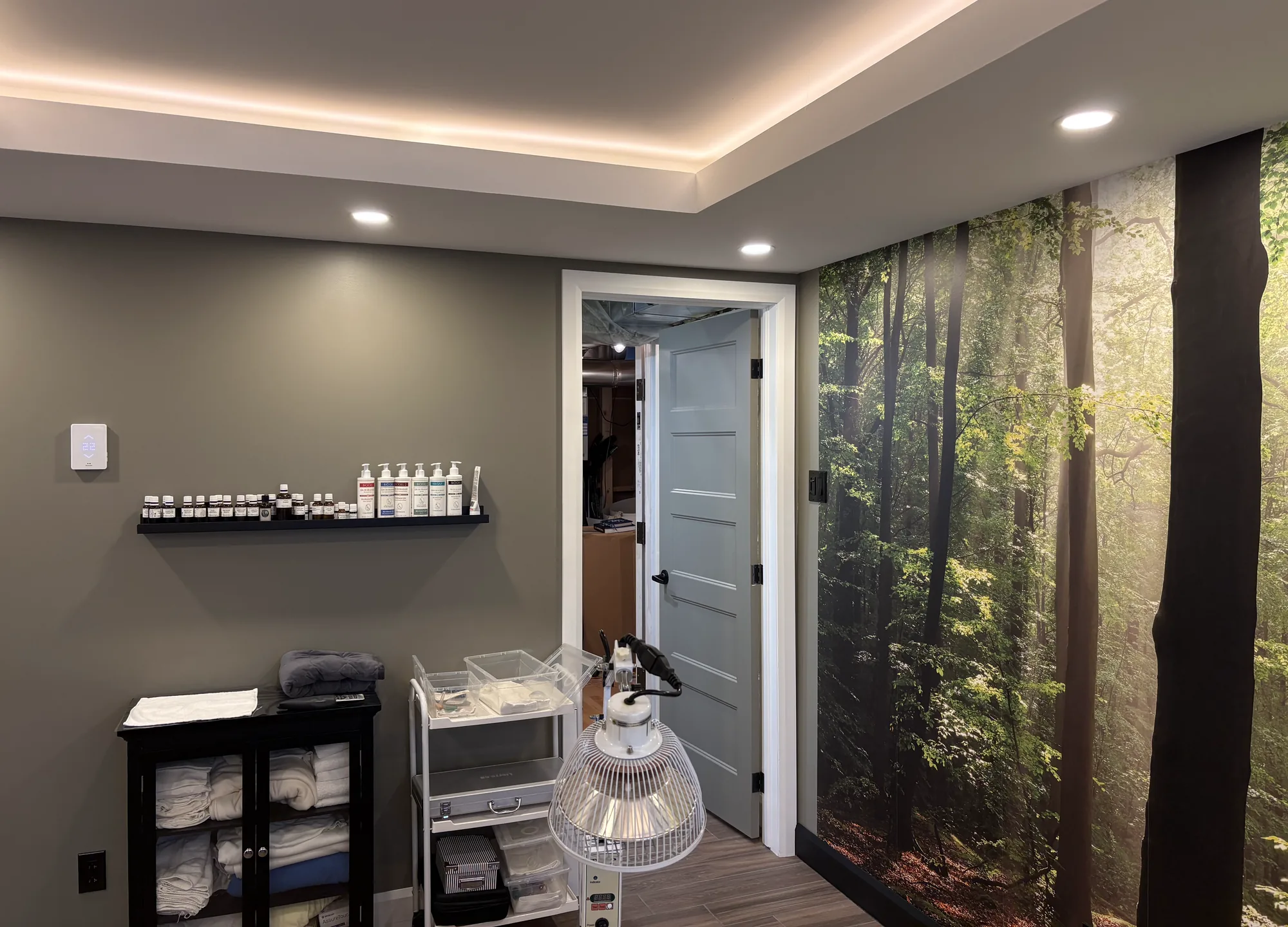 Salle de massothérapie : mur kaki avec une tablette de produits, porte bleu-gris ouverte, lampe infrarouge sur pied et grande fresque de forêt sur le mur de droite